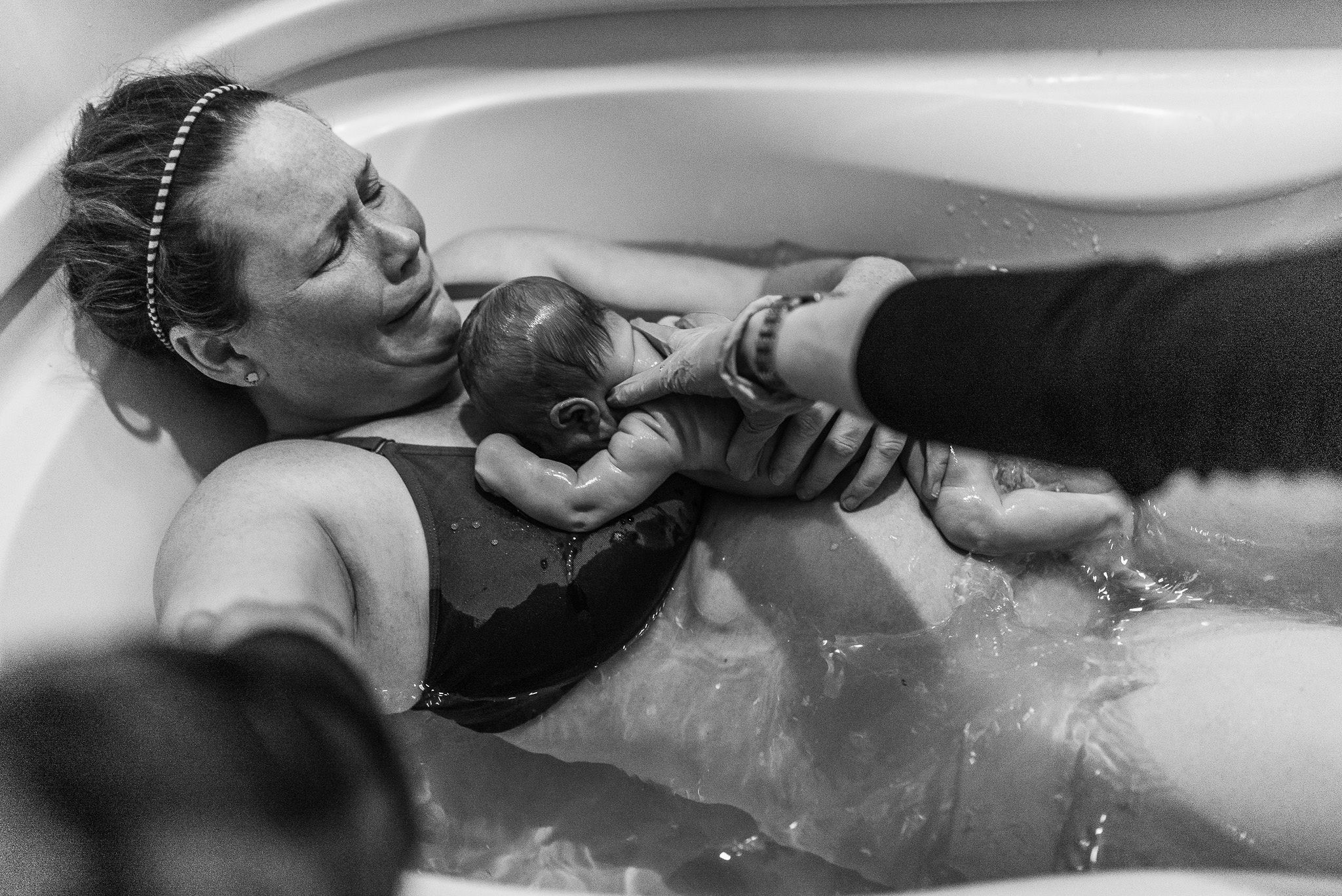 Mother holding her newborn in an inflatable birth pool while a caregiver supports the baby