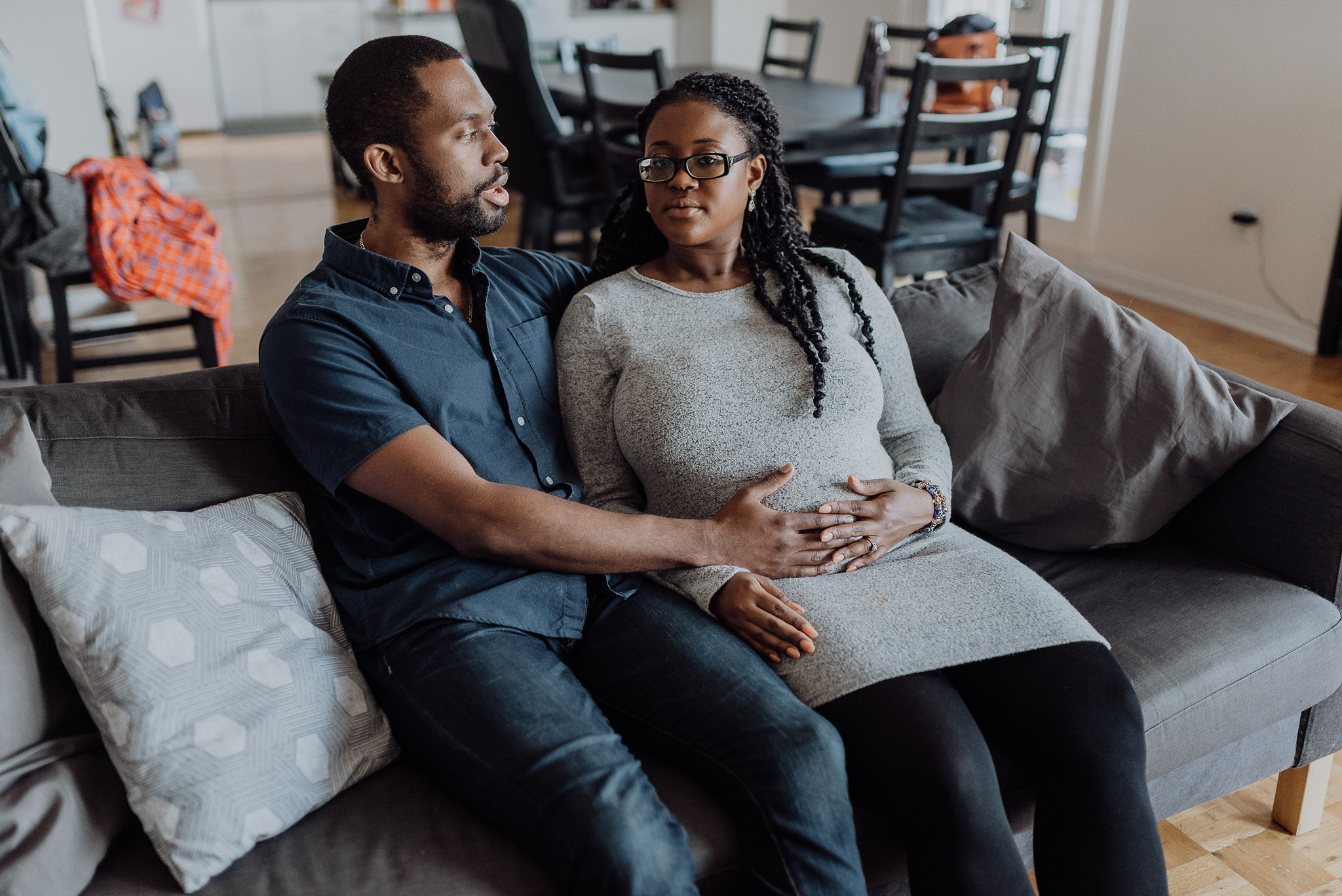 Expectant parents seated together on a sofa with hands resting on the pregnant parent's belly